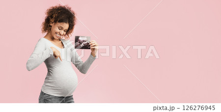 Look here. Excited afro pregnant woman with ultrasound scan pointing at tummy over pink background 126276945