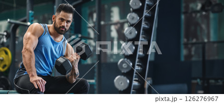 A man sits on a bench in a well-equipped gym, focusing on lifting a dumbbell with determination as he engages in his workout, panorama with copy space A man sits on a bench in a well-equipped gym, focusing on lifting a dumbbell with determination as he engages in his workout, panorama with copy space 126276967
