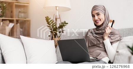 Shopping online is easy. Portrait of happy muslim woman in hijab with laptop and credit card sitting on sofa at home and smiling at camera Shopping online is easy. Portrait of happy muslim woman in hijab with laptop and credit card sitting on sofa at home and smiling at camera 126276968