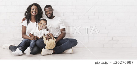 Family Bonds. Young African American Parents And Son Sitting On Floor Together, Looking At Camera Over White Brick Wall Background, Copy Space Family Bonds. Young African American Parents And Son Sitting On Floor Together, Looking At Camera Over White Brick Wall Background, Copy Space 126276997
