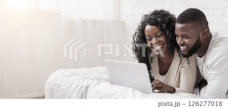 Happy african american couple using laptop while lying on bed together, browsing internet or watching movie 126277018