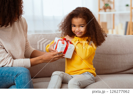Kid's Birthday. Cute African Little Girl Unpacking Wrapped Gift Sitting With Mother On Sofa At Home. 126277062