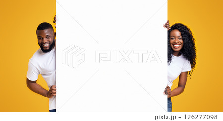 Black Girl And Guy Holding White Advertising Board, yellow studio background, panorama 126277098