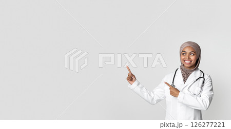 Medical Ad. Young black female therapist in hijab and white coat with stethoscope pointing at copy space over light background with her index fingers Medical Ad. Young black female therapist in hijab and white coat with stethoscope pointing at copy space over light background with her index fingers 126277221