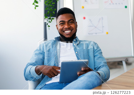 Cheerful african american business coach holding digital tab, sitting in modern office, looking at camera. 126277223
