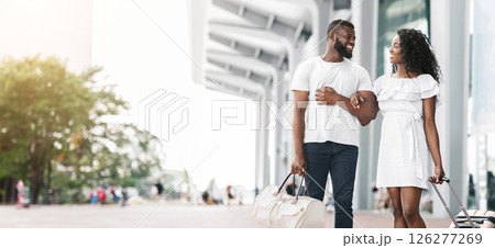 A smiling African American couple walks together outside a modern building on a sunny day. The man has his arm around the womans waist and they are both carrying luggage, copy space A smiling African American couple walks together outside a modern building on a sunny day. The man has his arm around the womans waist and they are both carrying luggage, copy space 126277269