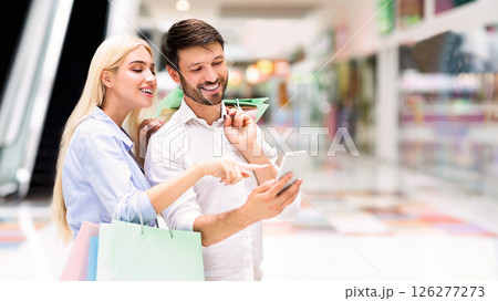 A couple is shopping together at the mall. They are looking at a smartphone, smiling and appearing to be happy. The woman is holding shopping bags and pointing at the screen of the smartphone 126277273
