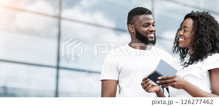 Black man and woman, dressed in casual clothing, stand outdoors and smile at each other while looking at documents. The couple is likely preparing for a trip, copy space Black man and woman, dressed in casual clothing, stand outdoors and smile at each other while looking at documents. The couple is likely preparing for a trip, copy space 126277462