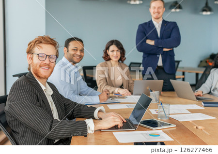 A group of four business professionals are working together in a modern office setting. One man is seated at a desk and working on a laptop, while another man and a woman are sitting beside him A group of four business professionals are working together in a modern office setting. One man is seated at a desk and working on a laptop, while another man and a woman are sitting beside him 126277466