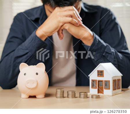 Worried man with a piggy bank, coin stacks, and house model nearby. Represents financial risks, property problems, money struggles, and the emotional toll of budgeting and unhappy banking situations. Worried man with a piggy bank, coin stacks, and house model nearby. Represents financial risks, property problems, money struggles, and the emotional toll of budgeting and unhappy banking situations. 126278350