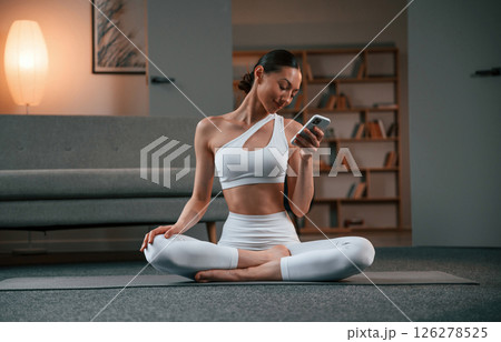 Holding smartphone. Young woman with slim body type in white sportswear is indoors doing yoga 126278525