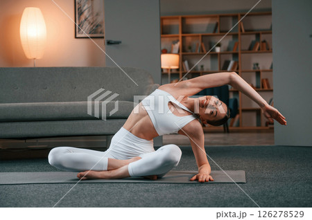 Stretching exercise. Young woman with slim body type in white sportswear is indoors doing yoga Stretching exercise. Young woman with slim body type in white sportswear is indoors doing yoga 126278529
