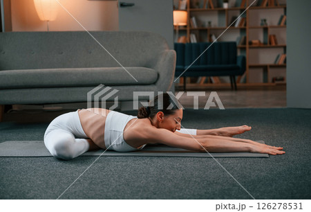 Stretching exercise. Young woman with slim body type in white sportswear is indoors doing yoga 126278531