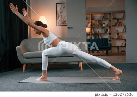 Warrior pose. Young woman with slim body type in white sportswear is indoors doing yoga 126278536