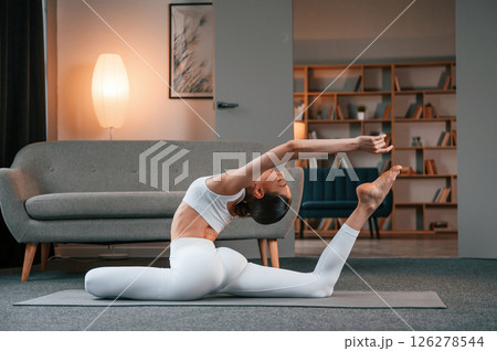 Practicing on the floor. Young woman with slim body type in white sportswear is indoors doing yoga Practicing on the floor. Young woman with slim body type in white sportswear is indoors doing yoga 126278544