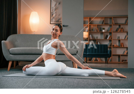 Practicing on the floor. Young woman with slim body type in white sportswear is indoors doing yoga 126278545