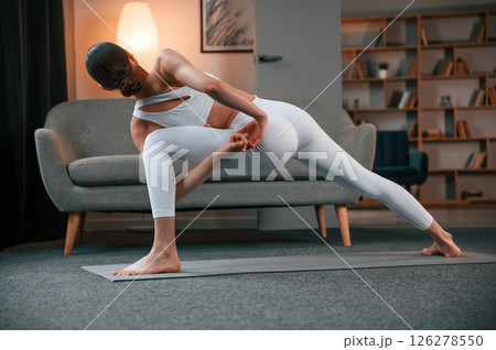 Warrior pose. Young woman with slim body type in white sportswear is indoors doing yoga Warrior pose. Young woman with slim body type in white sportswear is indoors doing yoga 126278550