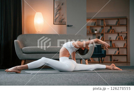 Practicing on the floor. Young woman with slim body type in white sportswear is indoors doing yoga 126278555