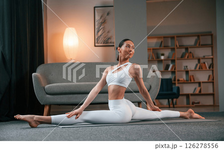 Practicing on the floor. Young woman with slim body type in white sportswear is indoors doing yoga Practicing on the floor. Young woman with slim body type in white sportswear is indoors doing yoga 126278556