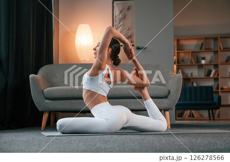 Professional yoga practitioner. Young woman with slim body type in white sportswear is indoors Professional yoga practitioner. Young woman with slim body type in white sportswear is indoors 126278566