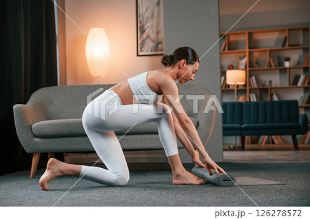 Lays out a yoga mat on the floor. Young woman with slim body type in white sportswear is indoors 126278572