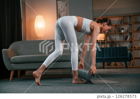 Lays out a yoga mat on the floor. Young woman with slim body type in white sportswear is indoors 126278573