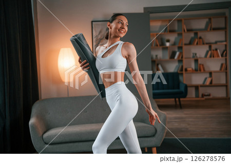 Standing and smiling. Young woman with slim body type in white sportswear is indoors with yoga mat Standing and smiling. Young woman with slim body type in white sportswear is indoors with yoga mat 126278576