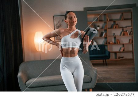 Standing and smiling. Young woman with slim body type in white sportswear is indoors with yoga mat 126278577