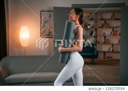 Woman with slim body type in white sportswear is indoors with yoga mat 126278584