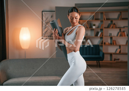 Ready for the session. Standing and holding yoga mat. Young woman with slim body type in white sportswear 126278585