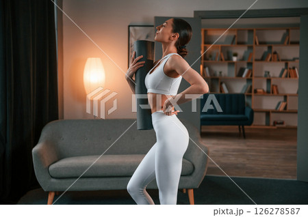 Ready for the session. Standing and holding yoga mat. Young woman with slim body type in white sportswear Ready for the session. Standing and holding yoga mat. Young woman with slim body type in white sportswear 126278587