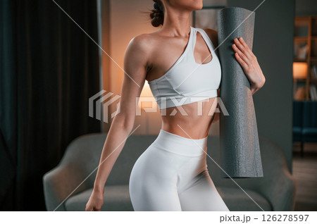 Close up view. Young woman with slim body type in white sportswear is indoors with yoga mat 126278597