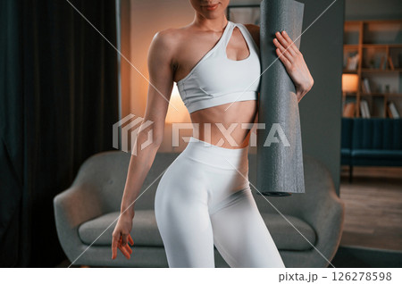 Close up view. Young woman with slim body type in white sportswear is indoors with yoga mat 126278598