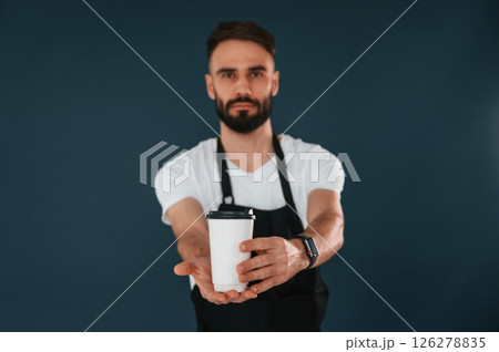 Giving the cup of coffee. Cafe worker is in apron. Handsome man is in the studio against blue background 126278835