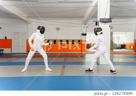 Two female fencers competing in bright indoor fencing hall. 126278990