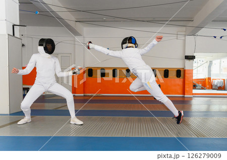 Female fencer making airborne lunge attack in indoor duel. 126279009