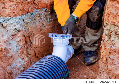 Construction worker adjusts drainage PVC pipe in newly excavated trench, ensuring precise during installation. Construction worker adjusts drainage PVC pipe in newly excavated trench, ensuring precise during installation. 126282144