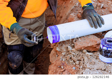 Construction worker is using blue adhesive on white PVC pipe on set up drainage pipe system in trench 126282146