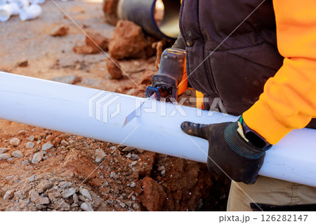 Skilled worker uses hand saw to cut plastic pipe at construction site, surrounded by materials. Skilled worker uses hand saw to cut plastic pipe at construction site, surrounded by materials. 126282147