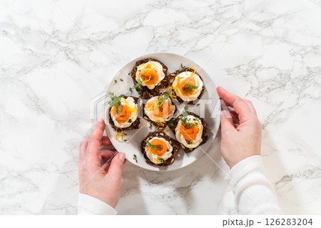 A top down view of a person holding a plate of smoked salmon canapes arranged on gourmet crispbread topped with cream cheese fresh herbs and lemon zest. The elegant appetizer is beautifully presented 126283204