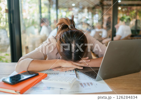 Tired woman resting her head on a desk surrounded by a laptop, documents, and a smartphone in a sunlit office. Tired woman resting her head on a desk surrounded by a laptop, documents, and a smartphone in a sunlit office. 126284834