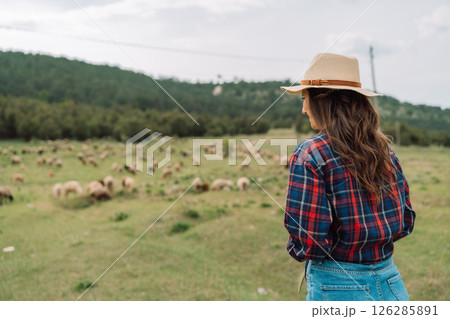 A woman wearing a straw hat stands in a field of sheep 126285891