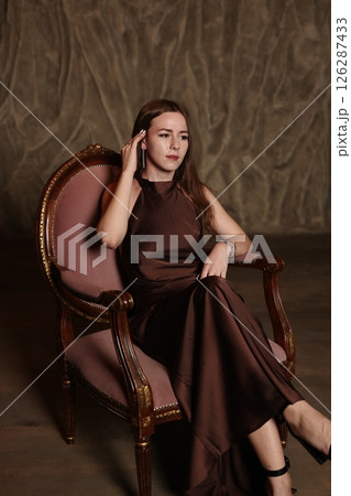 An Elegant Woman Dressed in a Beautiful Brown Dress Posing Gracefully on a Vintage Chair An Elegant Woman Dressed in a Beautiful Brown Dress Posing Gracefully on a Vintage Chair 126287433