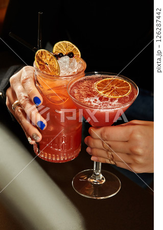 Women holding tropical tiki cocktails with dehydrated orange slices and ice cubes 126287442
