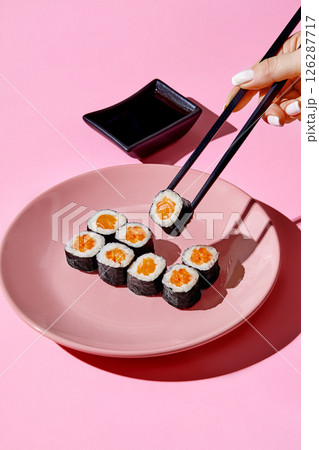 Woman using black chopsticks picking up salmon uramaki sushi roll from pink plate on pink background 126287717