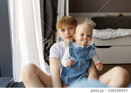 Older brother with baby sister in cozy home interior by window. Natural light, cute baby in blue dress 126290055