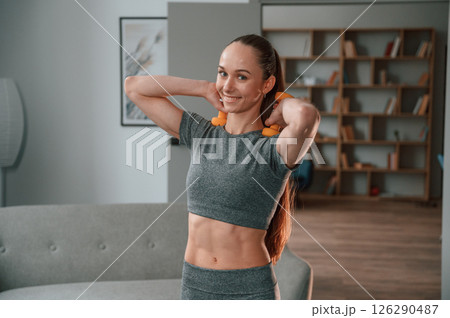 Smiling and holding dumbbells. Young woman in yoga clothes doing fitness indoors 126290487