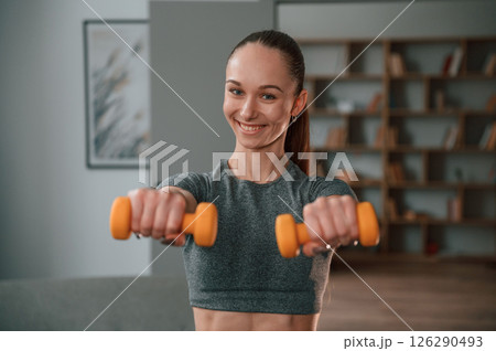 Positive young woman in yoga clothes is smiling and holding dumbbells 126290493