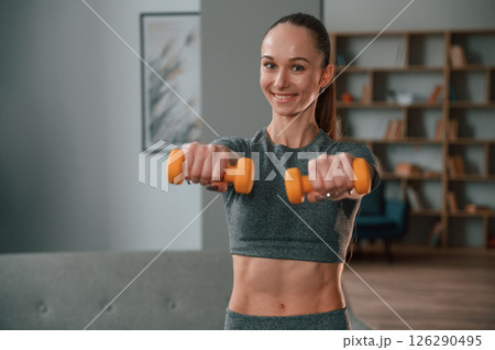 Positive young woman in yoga clothes is smiling and holding dumbbells Positive young woman in yoga clothes is smiling and holding dumbbells 126290495