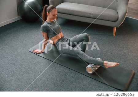 Legs exercises by using foam roller. Young woman in yoga clothes doing fitness indoors Legs exercises by using foam roller. Young woman in yoga clothes doing fitness indoors 126290558